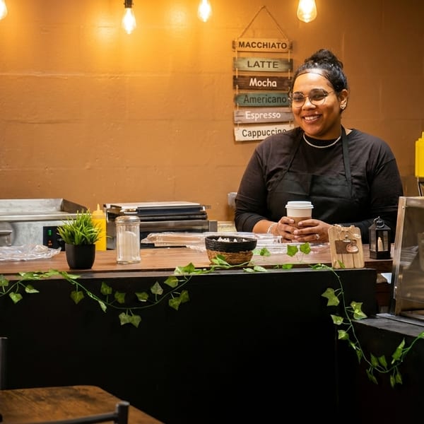 Smiling barista in an apron holding a coffee cup behind a counter with a menu board.