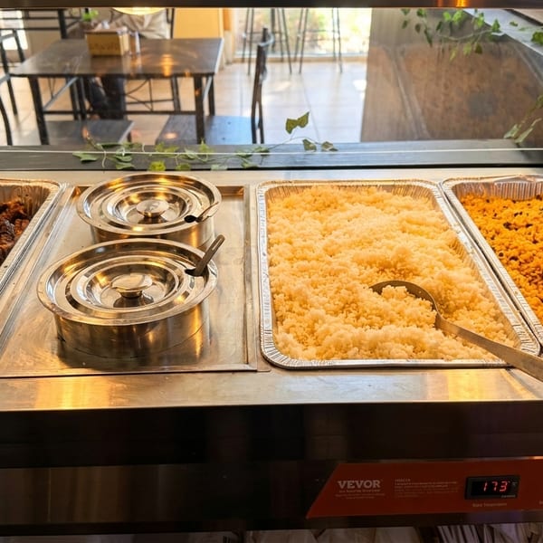 Buffet line with trays of chicken, white rice, and yellow rice, with a dining area in the background.