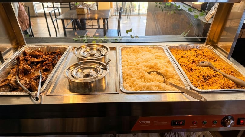 Buffet line with trays of chicken, white rice, and yellow rice, with a dining area in the background.